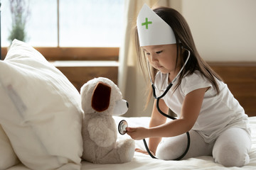 Asian little girl wear medical cap uniform hold phonendoscope listens heartbeat to stuffed toy dog...