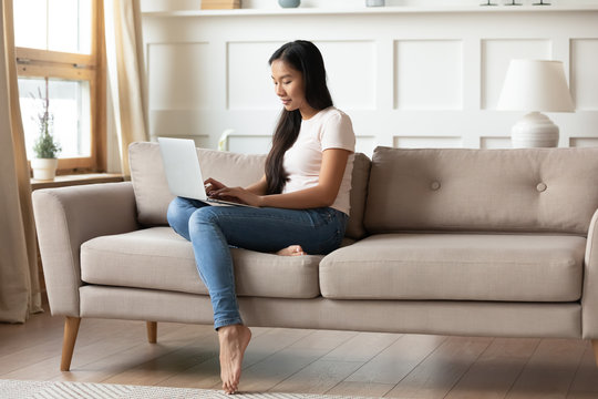 In modern living room Vietnamese young woman using laptop seated on sofa. Typing e-mail do remote work, chatting with friend boyfriend. Web surfing, booking hotels for future vacation holidays concept