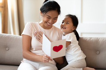 Asian lovely sweet preschool daughter congratulates mommy with life event birthday holiday, prepares for her handmade post card with red painted heart symbol of unconditional love, Mother Day concept