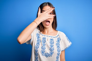 Young beautiful brunette woman wearing casual t-shirt standing over blue background peeking in shock covering face and eyes with hand, looking through fingers with embarrassed expression.