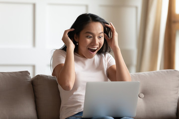 Crazy excited asian woman sit on couch read news online on laptop feels shocked amazed and...
