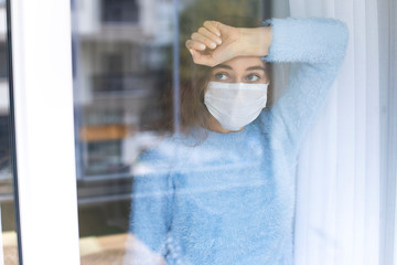 Young woman in quarantine wearing a mask and looking through the window - obrazy, fototapety, plakaty