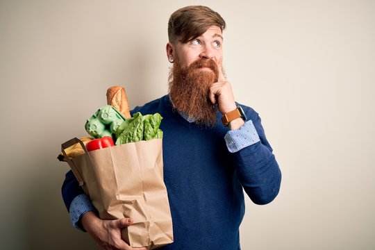 Redhead Irish man with beard holding fresh groceries from supermarkt over isolated background serious face thinking about question, very confused idea
