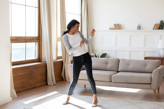 Joyful Black Young Woman Dancing In Bright Modern Living Room. Happy Girl Have Fun At Home On Lazy Weekend, Receive Good Or Pleasant News, Celebrate Moving To New Apartment Or House