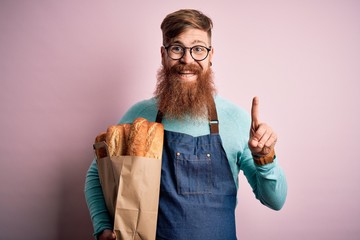 Irish redhead baker man with beard holding groceries paper bag of bread over yellow background...
