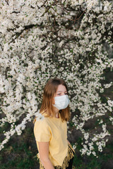 portrait of sad young woman in protective medical face mask with flowers near blooming tree in spring time. Coronavirus protection. epidemic of coronavirus. the aroma a tree in the garden on a spring