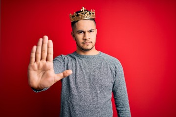 Young handsome man wearing golden crown of prince over isolated red background doing stop sing with...