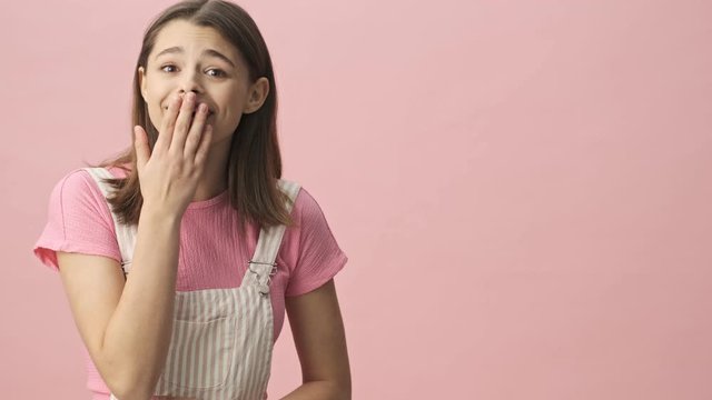 Surprised smiling pretty brunette woman in overalls pointing on copyspace and looking at the camera over pink background