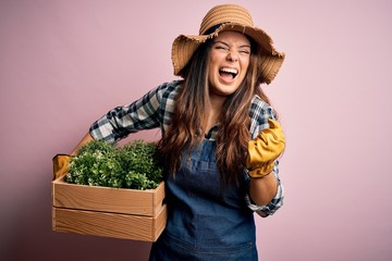 Young beautiful brunette farmer woman wearing apron and hat holding box with plants screaming proud...