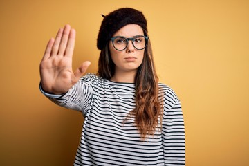 Young beautiful brunette woman wearing french beret and glasses over yellow background doing stop sing with palm of the hand. Warning expression with negative and serious gesture on the face.