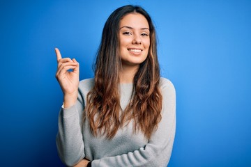 Young beautiful brunette woman wearing casual sweater standing over blue background with a big smile on face, pointing with hand and finger to the side looking at the camera.