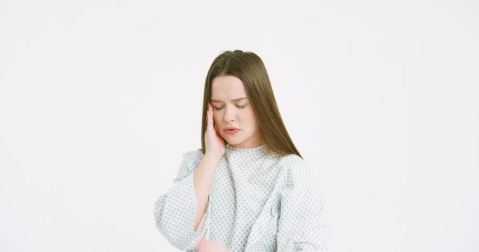 Camera zooming in at young Caucasian sick woman in night dress in hospital having severe headache. Female patient holding her forehead with fingers and feeling terrible pain on white wall background.