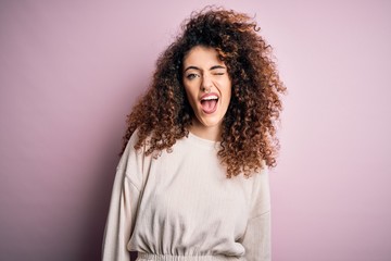 Beautiful woman with curly hair and piercing wearing casual sweater over pink background winking...