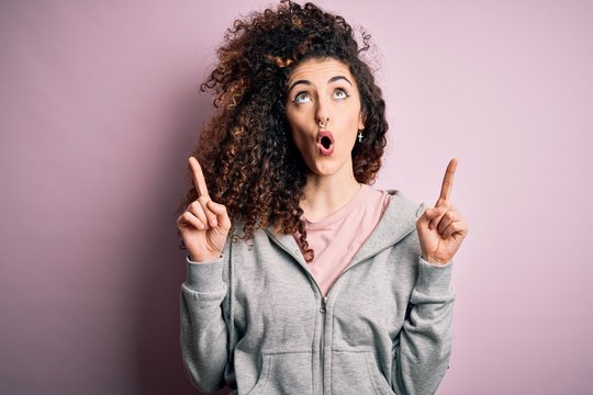 Young beautiful brunette sportswoman with curly hair and piercing wearing sportswear amazed and surprised looking up and pointing with fingers and raised arms.