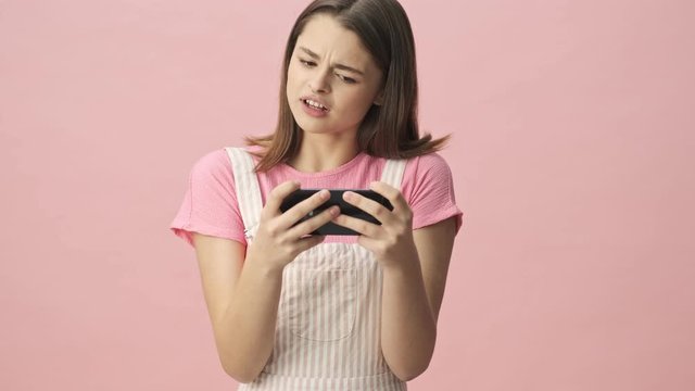 Confused pretty brunette woman in overalls playing on smartphone over pink background