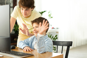 Smiling boy and teenager chatting online and waving at the computer screen. Quarantine and self-isolation due to coronavirus.