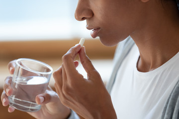 Close up of black girl hold glass of water and white pills at her mouth. Take painkillers,...