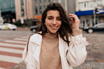 Obraz premium Close up outside portrait of smiling pretty woman with dark hair wearing beige sweater and white coat walking on the street with happy emotions on background of old buildings