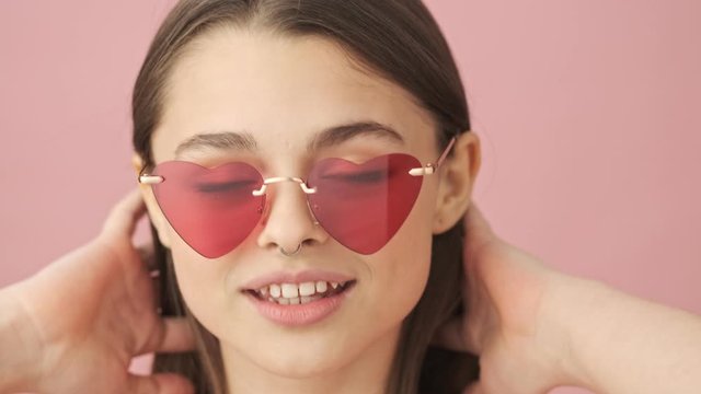 Close up view of happy pretty brunette woman in overalls and sunglasses posing and looking at the camera over pink background