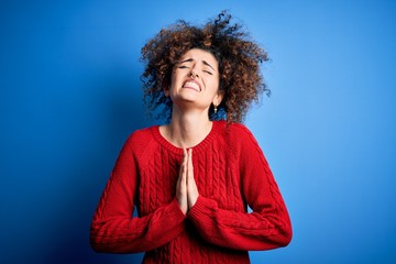 Young beautiful woman with curly hair and piercing wearing casual red sweater begging and praying...