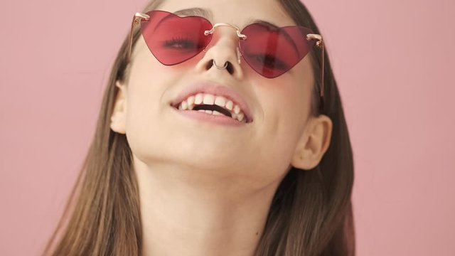 Close up view of smiling pretty brunette woman in overalls and sunglasses correcting her hairstyle and looking at the camera over pink background