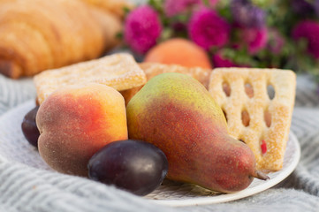 Warm summer spring picnic close-up with fresh pears and peaches on a gray plaid. Beautiful still life serving with sweet croissants and flowers. Good tasty mood background. Selective focus