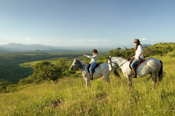 Two female horseback riders on horseback ride at sunset as one points overlooking the valley of...