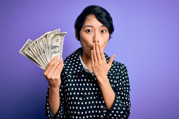 Young beautiful asian girl holding bunch of dollars banknotes over purple background cover mouth with hand shocked with shame for mistake, expression of fear, scared in silence, secret concept