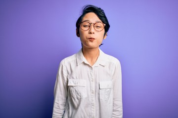 Fototapeta premium Young beautiful asian girl wearing casual shirt and glasses standing over purple background puffing cheeks with funny face. Mouth inflated with air, crazy expression.