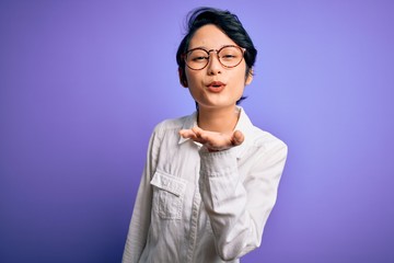 Young beautiful asian girl wearing casual shirt and glasses standing over purple background looking at the camera blowing a kiss with hand on air being lovely and sexy. Love expression.