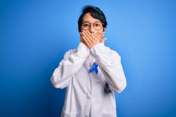 Young beautiful asian doctor girl wearing stethoscope and coat with blue cancer ribbon shocked covering mouth with hands for mistake. Secret concept.