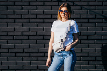Young model girl in white t-shirt and glasses with waist bag against a black brick wall