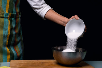 Working in the kitchen. pouring sweet sugar in bowl. Colorful apron. Cooking apple pie. Steel bowl with pieces of fruit. Cook table. Dark black background.