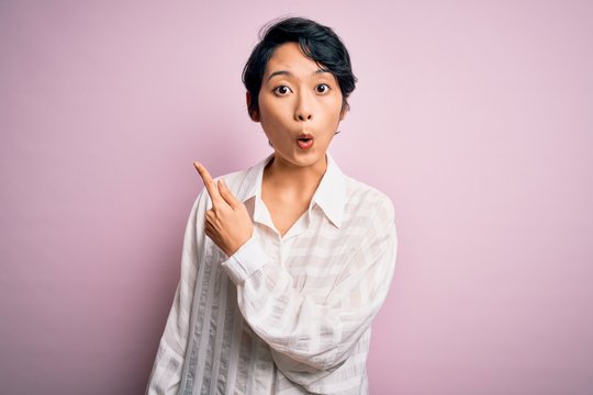 Young beautiful asian girl wearing casual shirt standing over isolated pink background Surprised pointing with finger to the side, open mouth amazed expression.