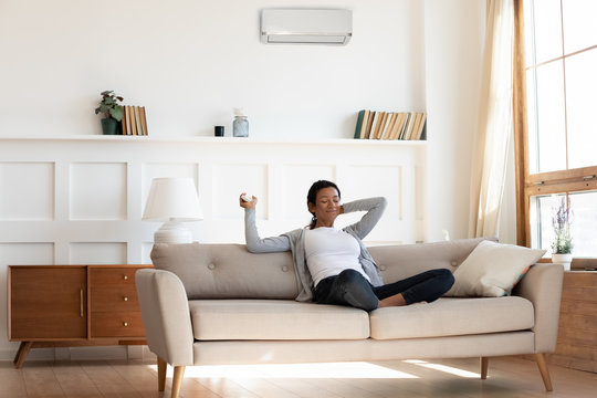 African American Millennial Girl, Contented And Calm, Sit On Comfortable Sofa In Cozy Modern Living Room And Hold Remote Control For The Air Conditioner. Enjoy Cool Fresh Air In Home