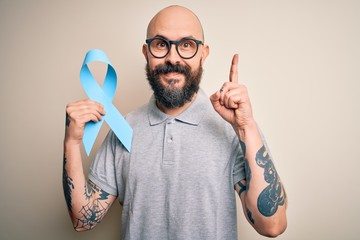 Handsome bald man with beard and tattoos holding blue cancer ribbon over isolated background...