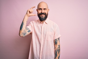 Handsome bald man with beard and tattoo wearing casual shirt over isolated pink background smiling and confident gesturing with hand doing small size sign with fingers looking and the camera. Measure