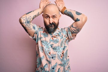 Handsome bald man with beard and tattoo wearing casual floral shirt over pink background Doing...