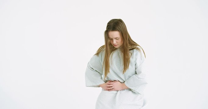 Caucasian sick female medical patient in hospital night dress having strong pain spasm in stomach on white wall background. Young woman holding her belly and feeling bad ache or nausea.