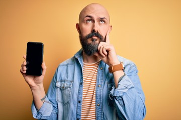 Handsome bald man with beard holding smartphone showing screen over yellow background serious face...