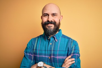 Handsome bald man with beard wearing casual shirt standing over isolated yellow background happy face smiling with crossed arms looking at the camera. Positive person.