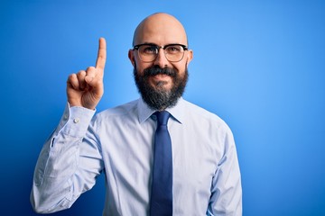 Handsome business bald man with beard wearing elegant tie and glasses over blue background showing and pointing up with finger number one while smiling confident and happy.
