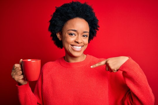 African American afro woman with curly hair drinking cup of coffee over red background with surprise face pointing finger to himself
