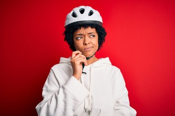 Young African American afro cyciling woman with curly hair wearing bike security helmet with hand on chin thinking about question, pensive expression. Smiling with thoughtful face. Doubt concept.