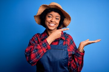 Young African American afro farmer woman with curly hair wearing apron and hat amazed and smiling to the camera while presenting with hand and pointing with finger.