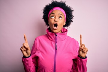 Young beautiful African American afro sportswoman with curly hair wearing pink sportswear amazed and surprised looking up and pointing with fingers and raised arms.