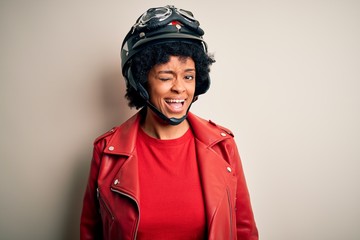 Young African American afro motorcyclist woman with curly hair wearing motorcycle helmet winking looking at the camera with sexy expression, cheerful and happy face.
