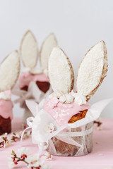 Fototapeta na wymiar Easter Cakes Kulich with Top Covered Pink Cream Decorated Cookies in Bunny Ears Form Copy Space. Christianity Traditional Festival Sweet Dessert in Textile Bag and Apricot Branch on Table