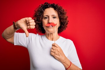 Middle age curly hair woman holding funny mustache over isolated red background with angry face, negative sign showing dislike with thumbs down, rejection concept