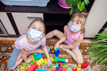 Two girls sisters in medical masks sit at home and play in a children's colorful constructor during isolation.